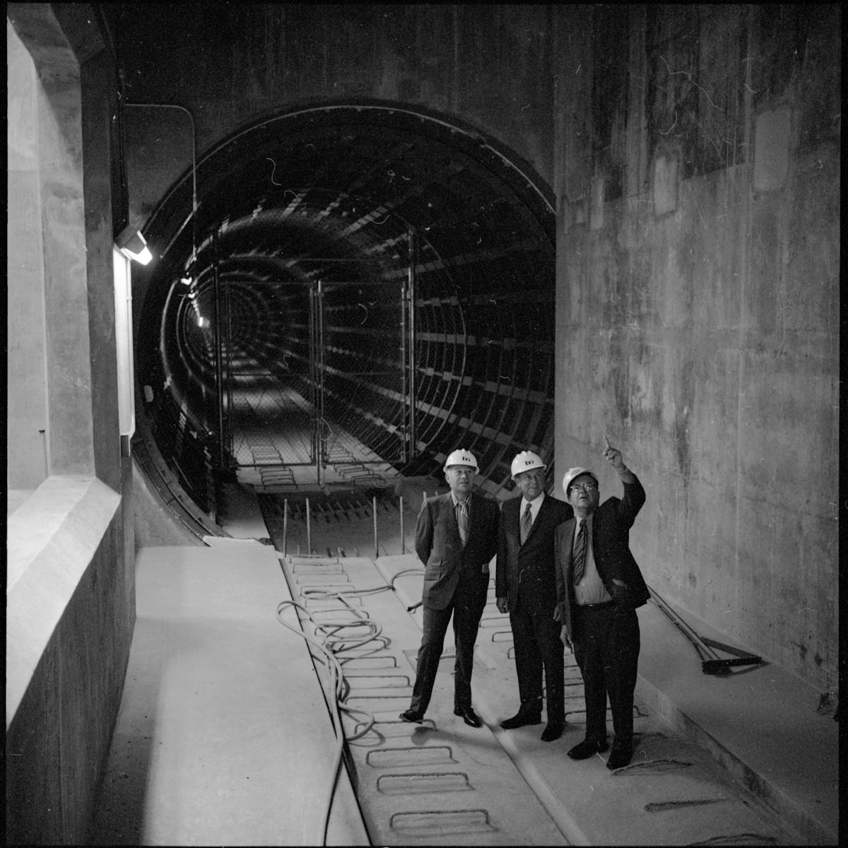 Three men in hard hats stand in a large circular tunnel under construction, circa 1970.