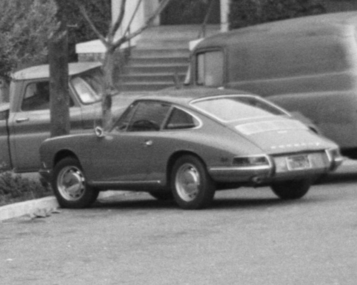Early Porsche 911 parked on street next to utility vehicles and pole, black and white photograph.