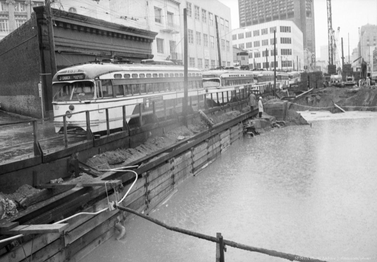 Vintage streetcar parked beside flooded BART tunnel construction site with urban buildings in background, 1971.