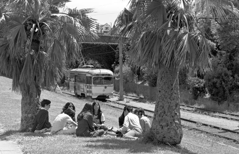 Group of young people sitting in circle under palm trees with vintage streetcar passing behind them.