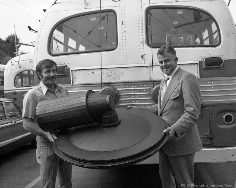 Two men display a Lockheel Kinetic Energy Wheel mockup in front of a transit bus, 1972.