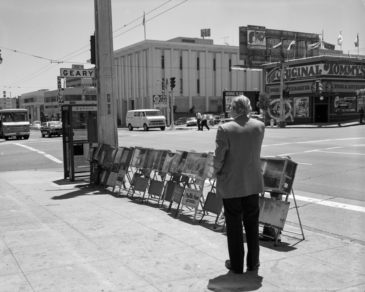 Man standing at bus stop with newspaper racks on Van Ness and Geary, San Francisco, 1974