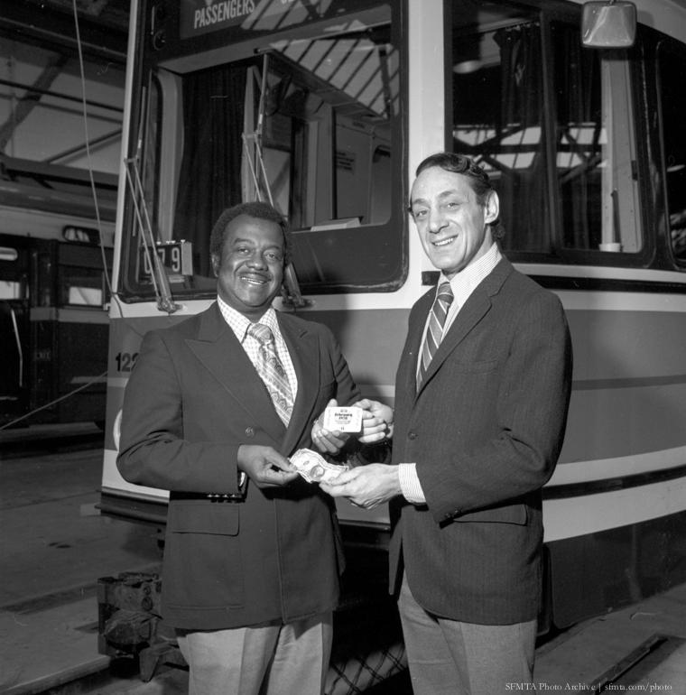 Two men in business suits exchanging transit passes in front of a Muni bus, 1978.