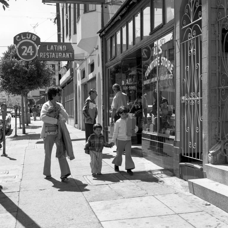 Family walking past storefronts on 24th Street Mission District sidewalk, 1978