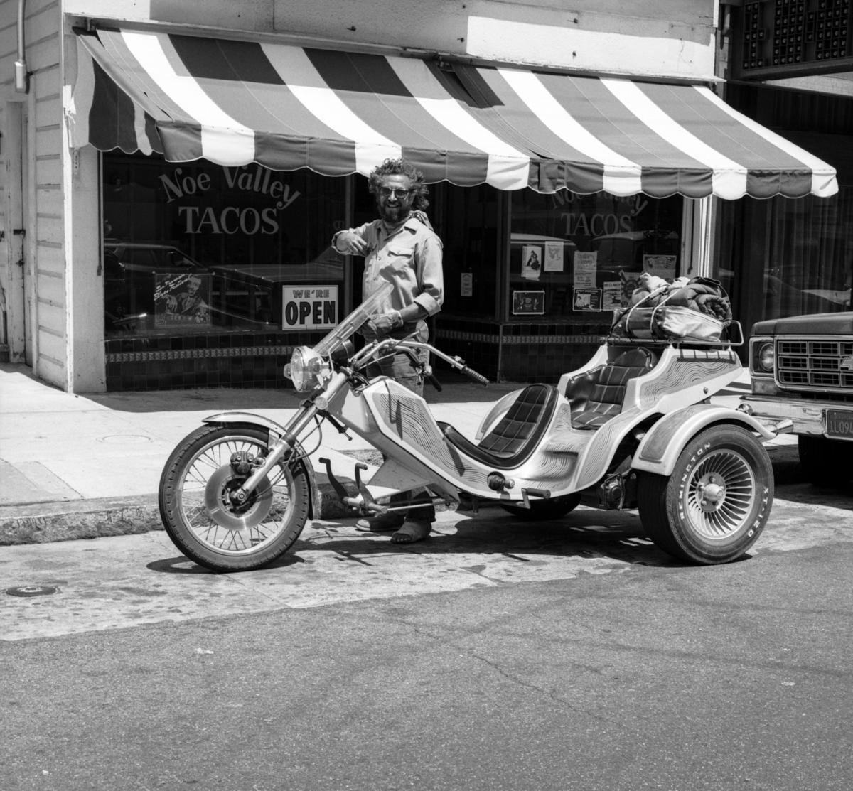 Man with custom motorcycle trike parked outside Noe Valley Tacos storefront, 1978.