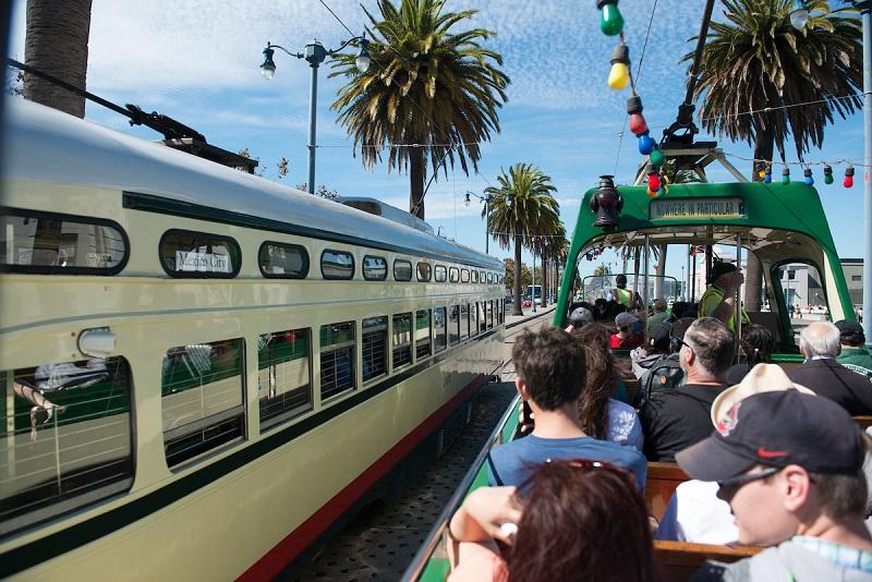 Passengers aboard a historic green and cream streetcar on The Embarcadero with palm trees and a modern train alongside.
