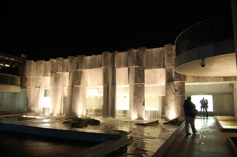 Illuminated stone waterfalls at night in an urban plaza with a visitor viewing the installation.