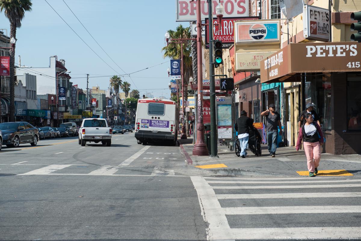 SFMTA bus stopped at Mission Street curb with pedestrians and storefronts visible
