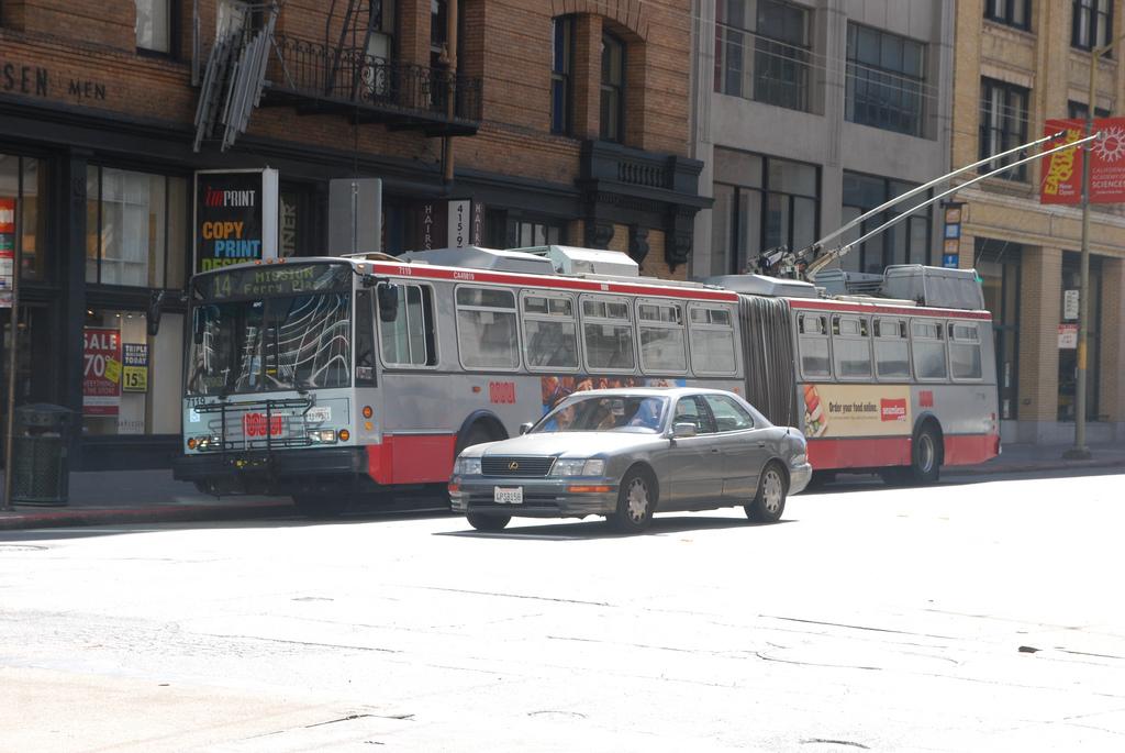Red and white Muni trolley bus stopped on Mission Street in downtown San Francisco with a silver sedan parked in front.