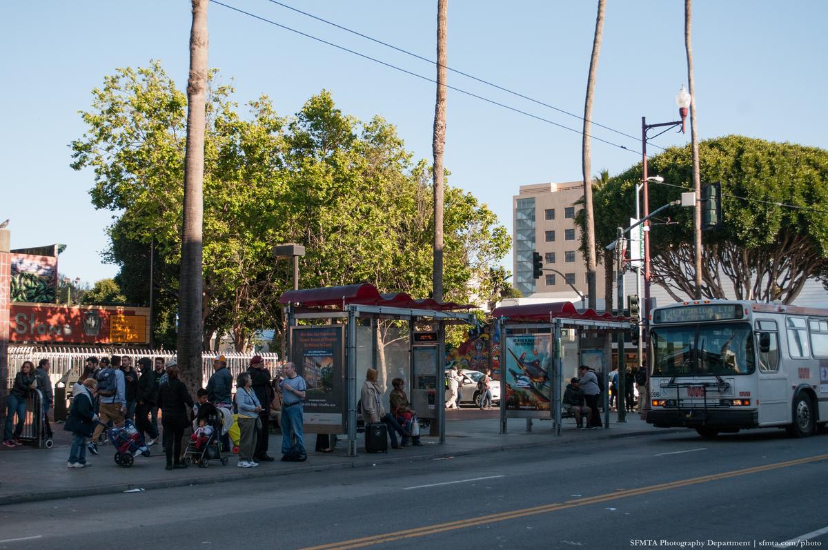 Muni bus at crowded 24th Street transit stop with passengers waiting under shelter.
