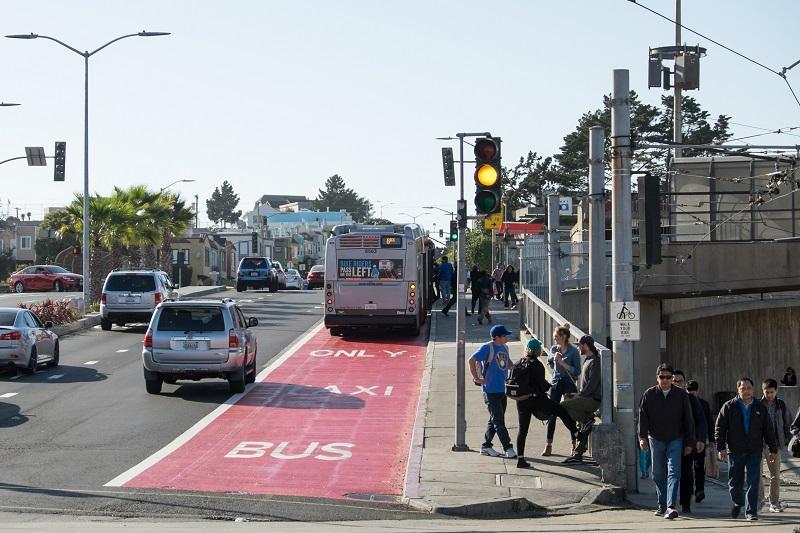 Bus pulling into red transit-only lane at Balboa Park Station with pedestrians waiting nearby and traffic passing.