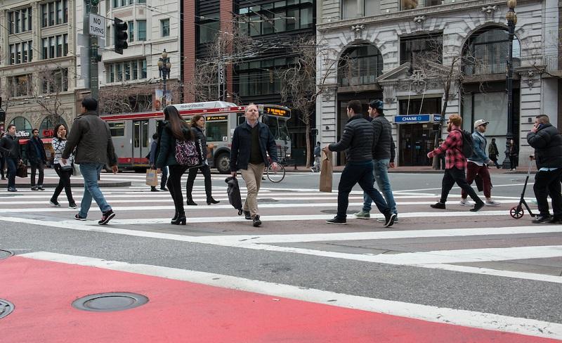 Pedestrians crossing at Market Street crosswalk with historic buildings and Muni bus in background.