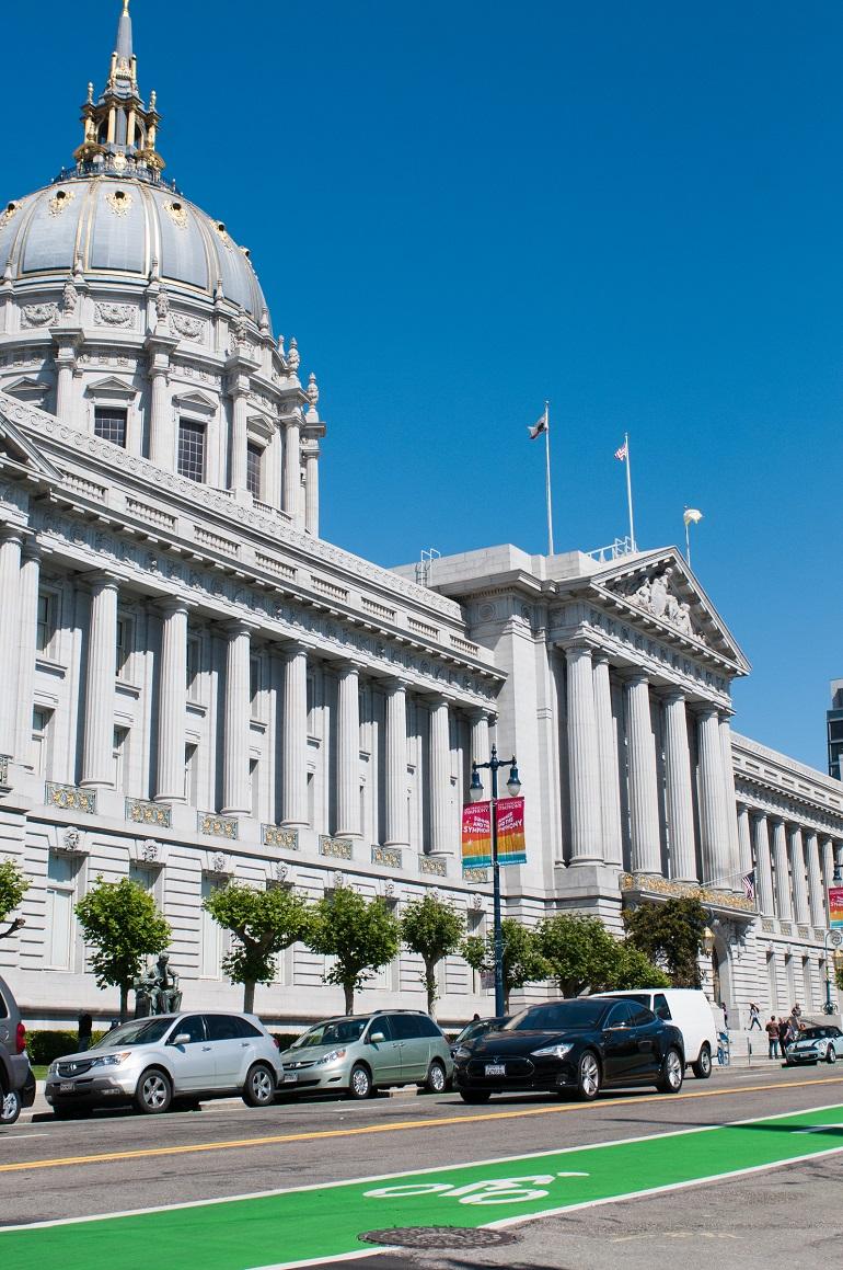 San Francisco City Hall with green raised bikeway in foreground and parked cars along street.