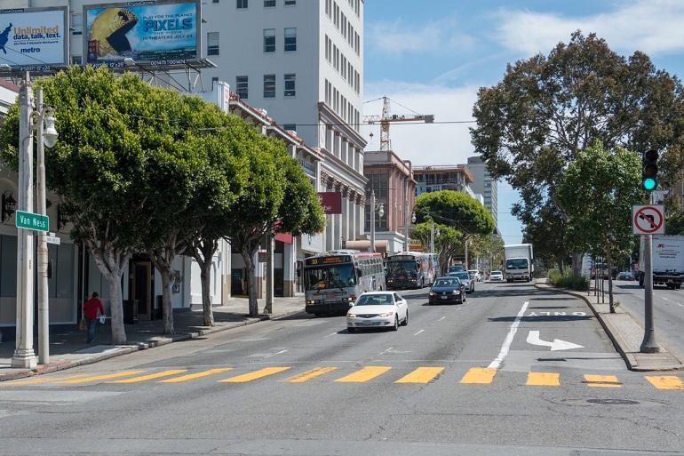 Muni bus traveling north on Van Ness Avenue with traffic and pedestrians on a tree-lined urban street.