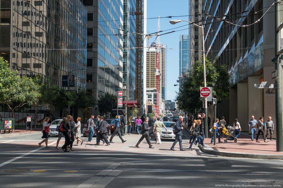Pedestrians crossing Market Street in downtown San Francisco with tall buildings and street infrastructure visible.