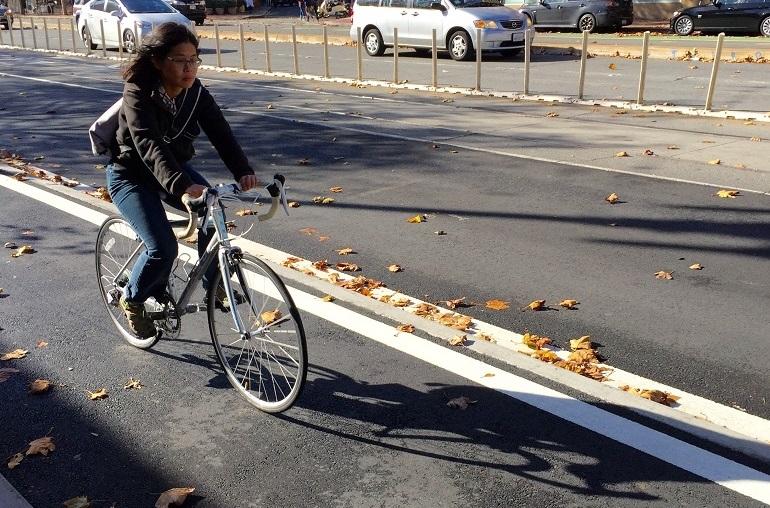Woman riding a bicycle on a raised bikeway with parked cars visible in the background.