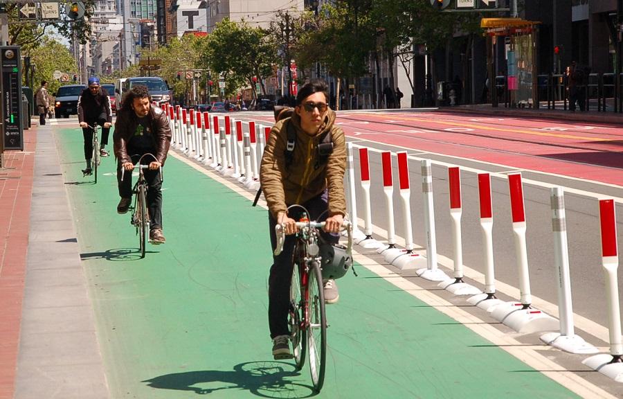 Cyclists riding on a green protected bike lane separated from traffic by white and red barrier posts on a city street.