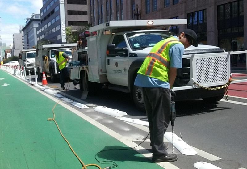 Workers in safety vests install protective bollards on a green transit lane using drilling equipment.