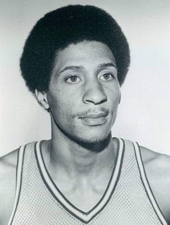 Black and white portrait photograph of a man wearing a mesh basketball jersey, circa 1975.