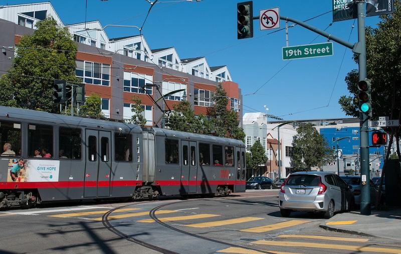 Red and gray Muni light rail train at 19th Street intersection with green traffic signal and silver car waiting.