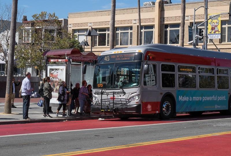 Passengers board a red and teal Muni bus at a street stop with historic buildings in the background.