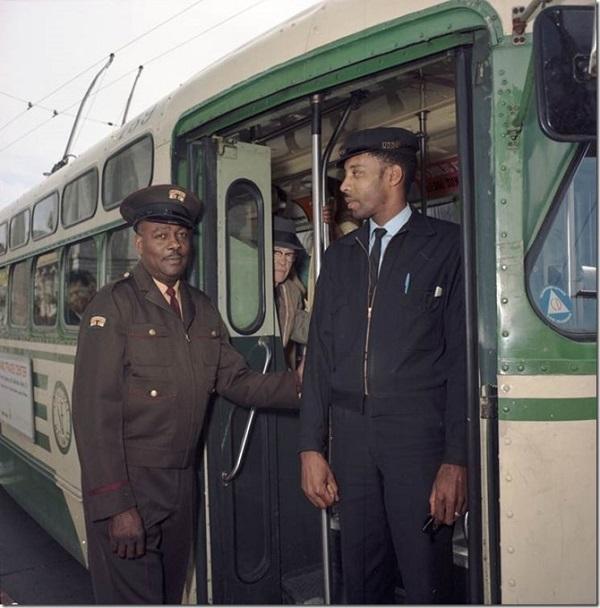 Bus operator in olive drab uniform standing at vehicle door next to man in dark suit, 1968.