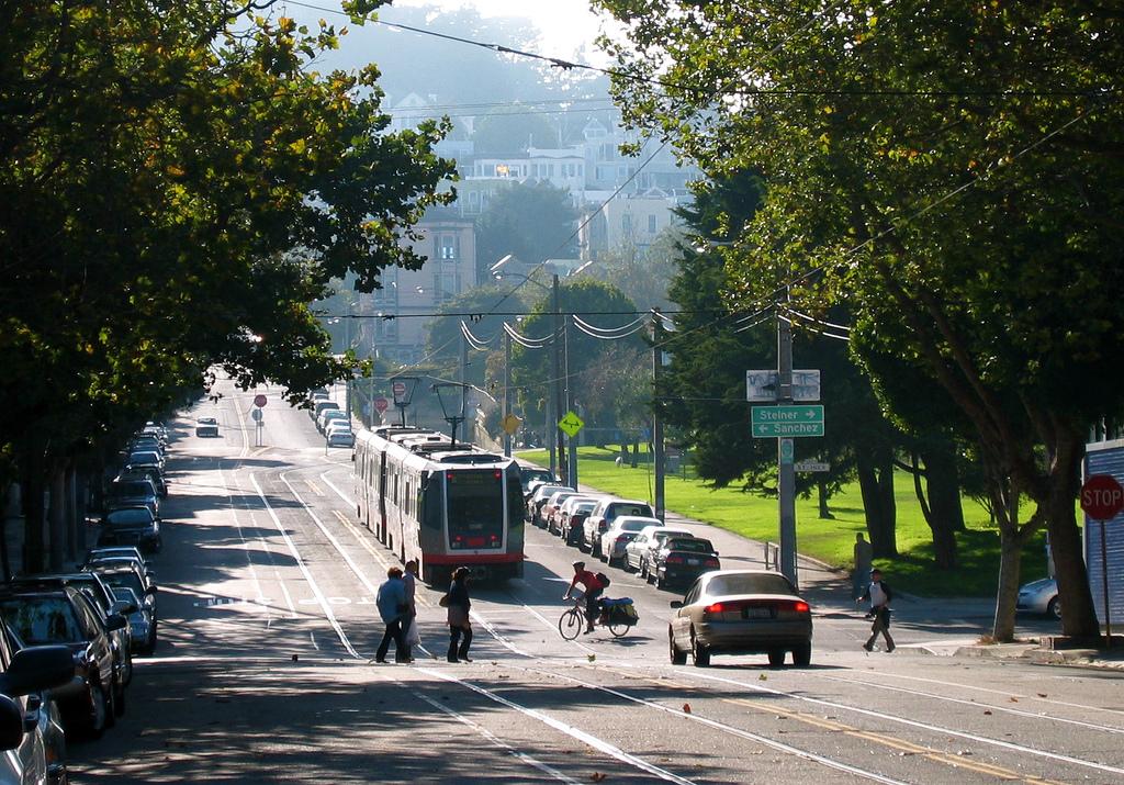 N Judah light rail vehicle traveling west on tree-lined Duboce Avenue with pedestrians and bicyclists nearby.