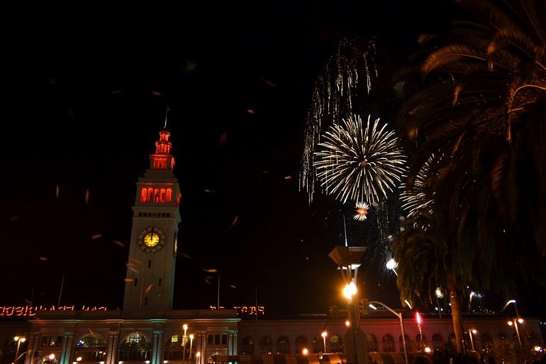 Ferry Building at night with fireworks exploding overhead and palm trees visible on the sides.