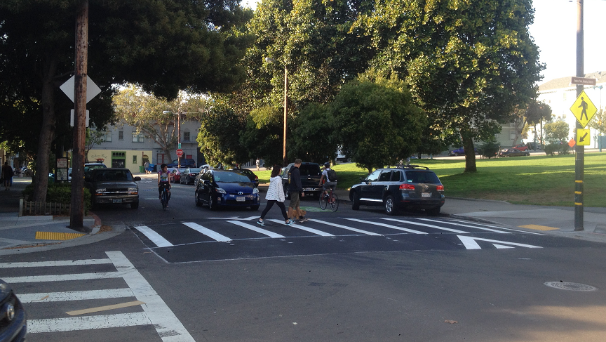 Pedestrians and cyclists crossing at a raised crosswalk intersection with parked and moving vehicles.