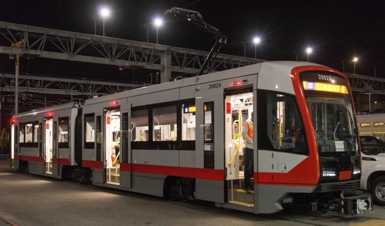 Red and gray light rail train stopped at night under overhead infrastructure and lighting at transit facility.