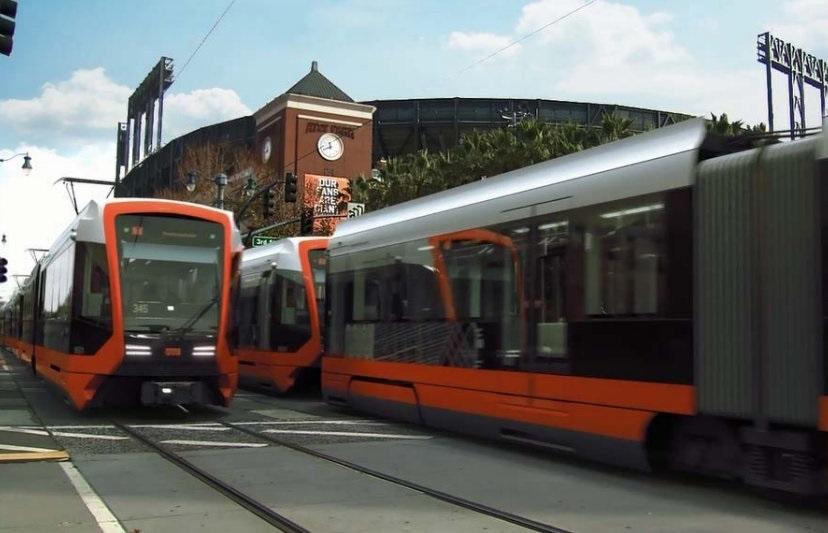 Red and silver light rail train on tracks with brick building and clock tower in background.