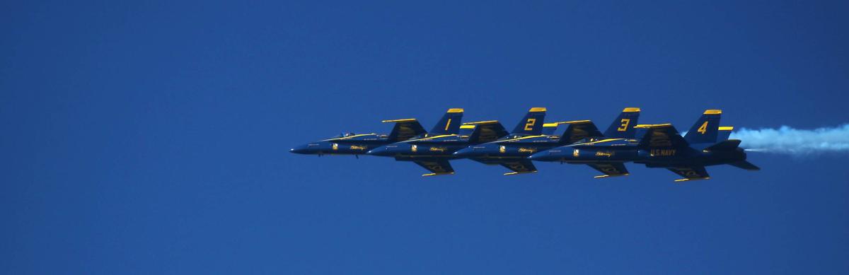 Blue Angels aircraft in formation flying through clear blue sky with smoke trails.