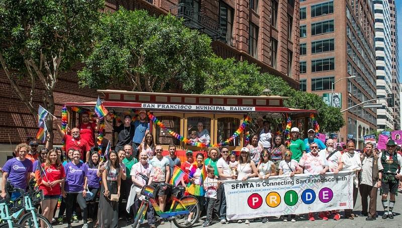 SFMTA staff and supporters on a decorated cable car at San Francisco Pride Parade, holding rainbow pride banner.