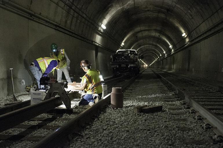 Workers in safety vests perform maintenance on railroad tracks inside a tunnel lit by overhead lights.