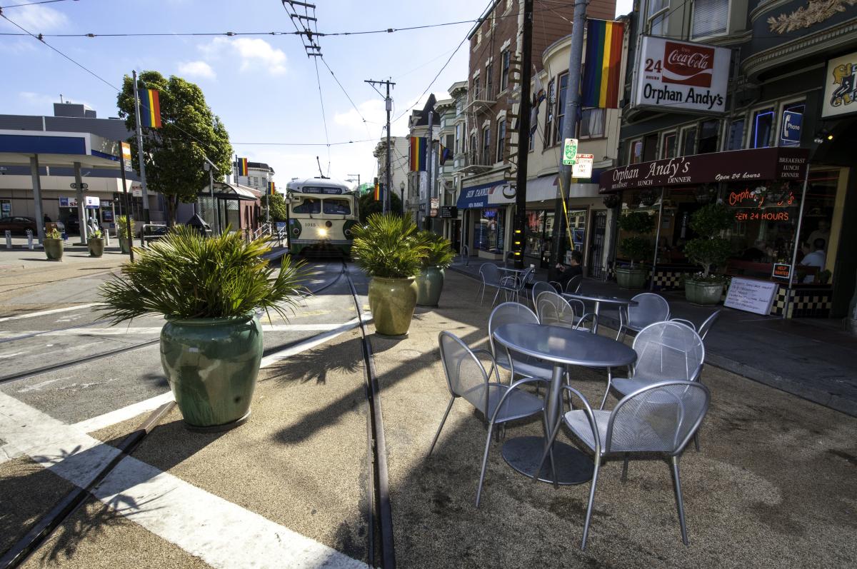 Outdoor seating area on Castro Street with metal chairs around a table, green planters with plants, and a historic Muni streetcar visible on the tracks.