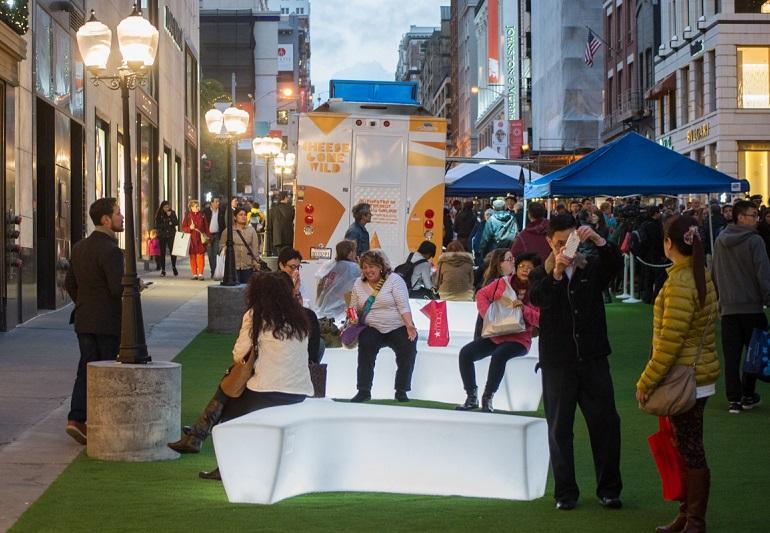 Crowds gather on a green-carpeted pedestrian promenade with white seating, blue tents, and illuminated storefronts along an urban street.