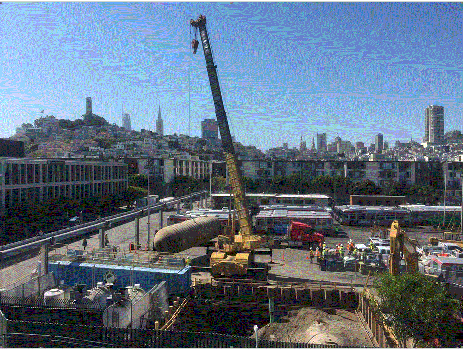 Construction crane removing large underground storage tank at urban transit facility with San Francisco skyline in background.