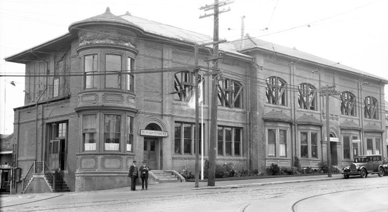 Historic brick building with rounded corner tower, large windows, and utility poles, circa 1928.