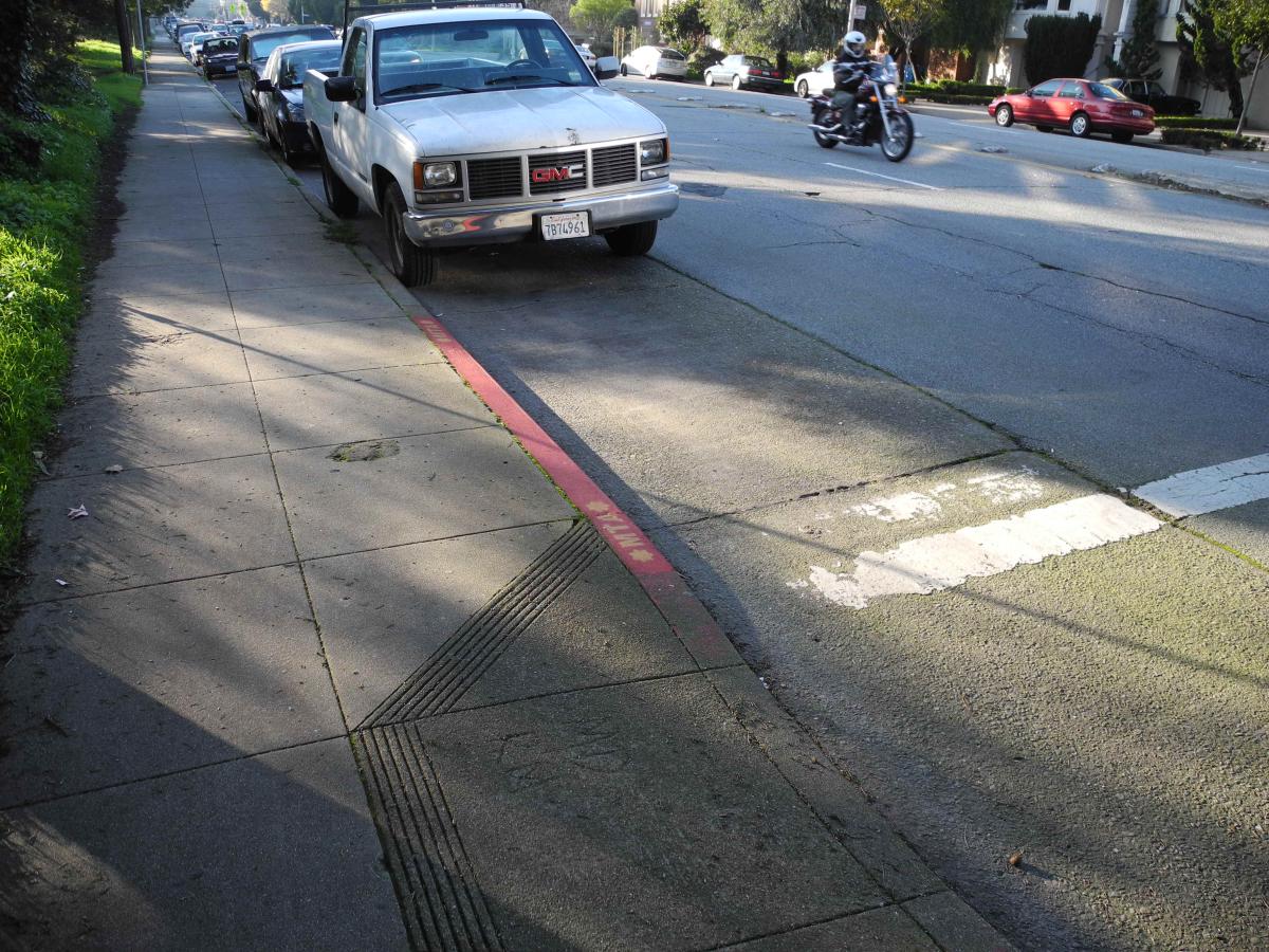 Red curb marking at street intersection improves pedestrian visibility near crosswalk and parked vehicles.
