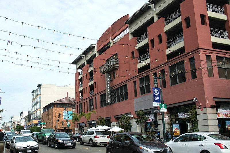 Red brick building with street-level retail, parked cars, and overhead string lights on urban street.