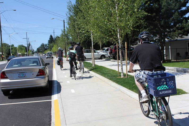 Cyclists ride on a protected bike lane separated from traffic by parked cars on a tree-lined street.