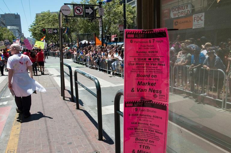 Bright pink alert sign posted at a Muni bus stop displaying detoured route information during Pride parade on Market Street.