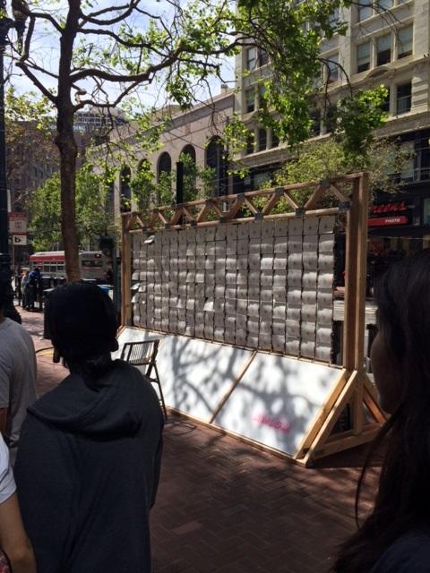 Wooden-framed installation with grid of white panels on urban street, pedestrians nearby, historic buildings in background.