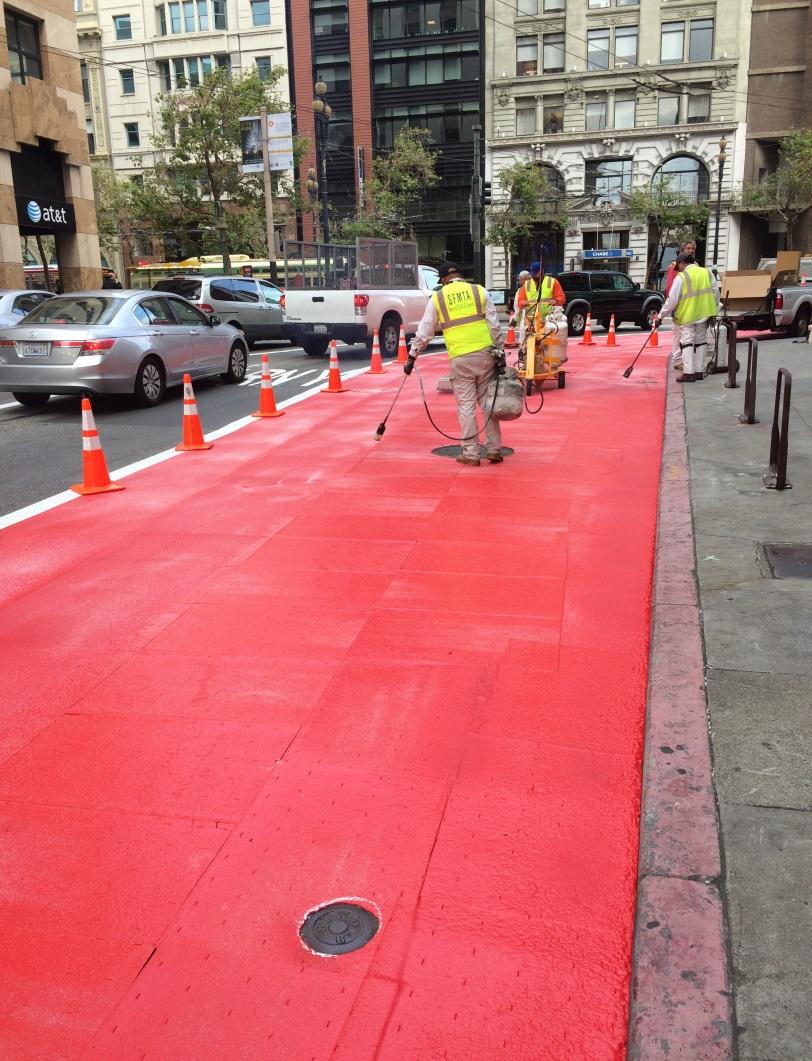 Workers in safety vests paint a red transit-only lane on a city street surrounded by buildings and traffic.