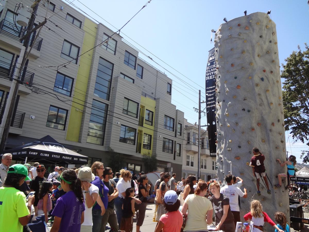 Crowd of people gathered around a tall rock climbing wall at an outdoor street event.