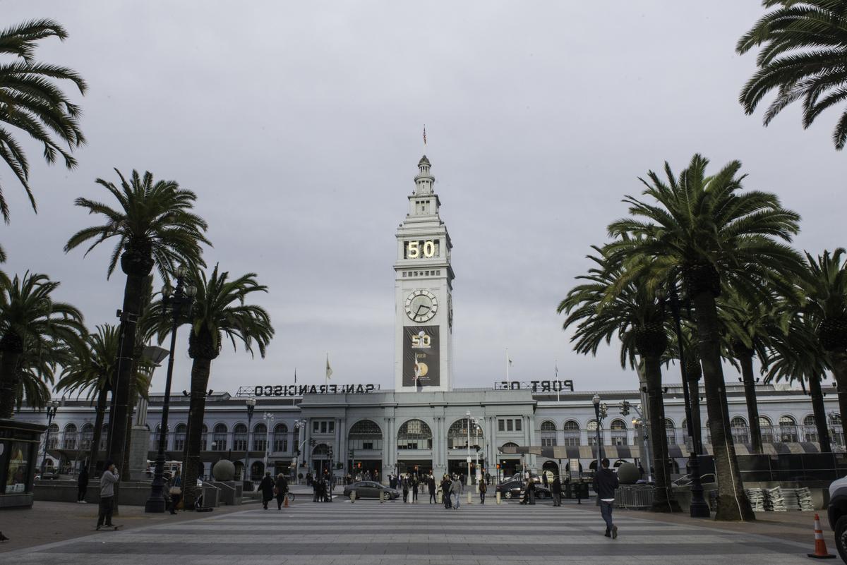 Ferry Building clock tower with palm trees and plaza, Super Bowl 50 banners visible on facade.