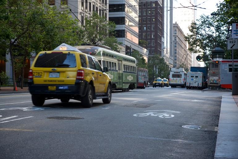 Yellow taxi and historic streetcar on busy Market Street with Muni buses and city buildings.