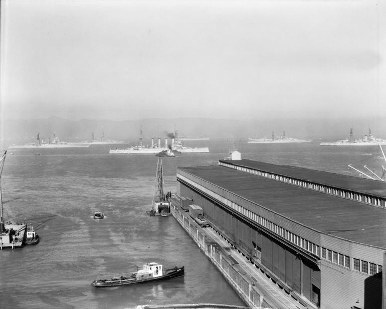 Black and white photograph of San Francisco Bay waterfront with multiple naval vessels anchored in the water and a pier structure in the foreground.