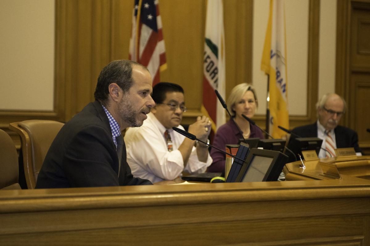 Four people seated at a panel table with microphones during a board meeting in City Hall, with American and California flags in the background.