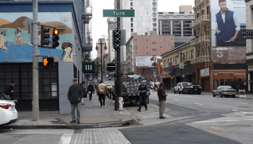 Pedestrians cross downtown streets at Turk Street intersection with traffic light and buildings visible.